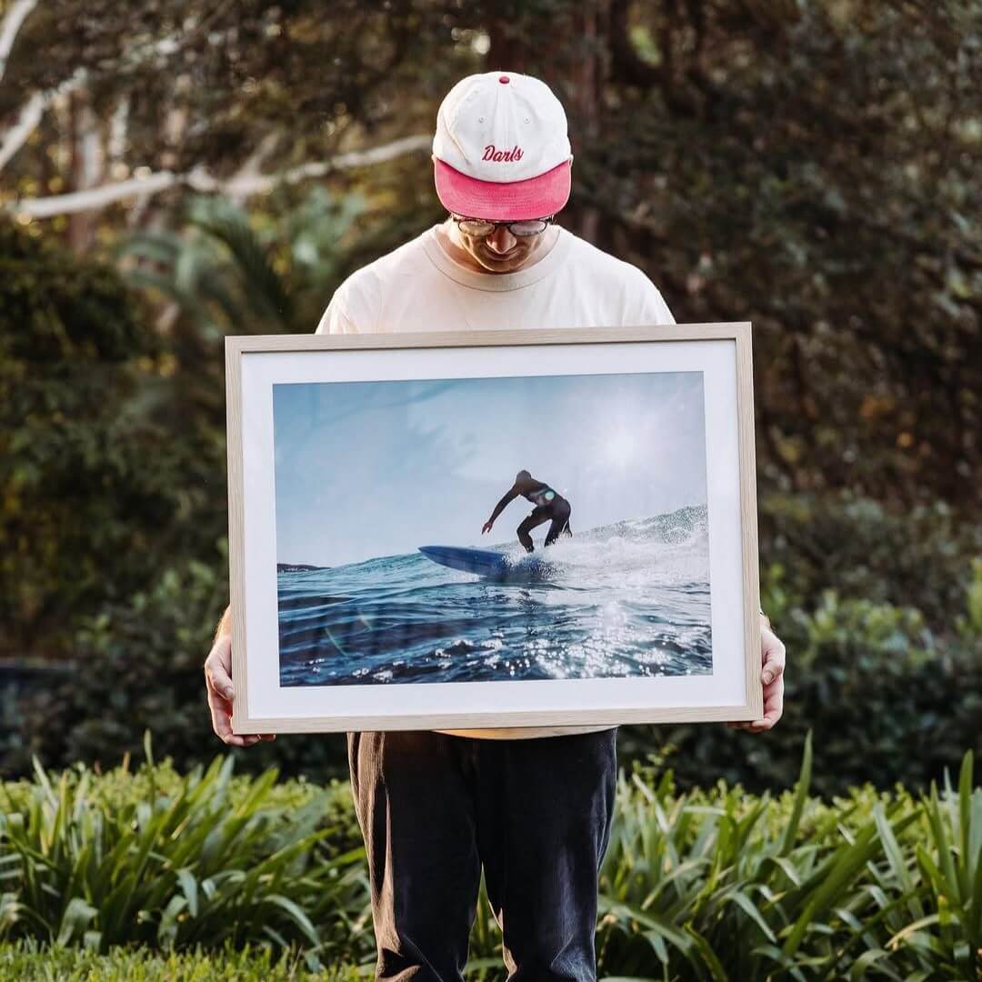 A man in a red and white cap stands holding and looking down at a framed photograph of a man catching a wave on a surfboard.